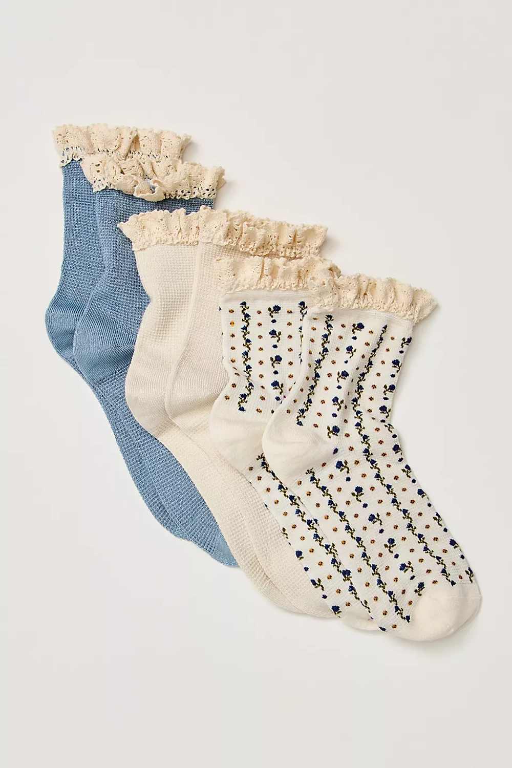Three pairs of women's ankle socks arranged diagonally on a light gray background. The set includes a dusty blue pair, an off-white pair with beige ruffles, and an off-white pair with a black and blue floral pattern and beige ruffles.