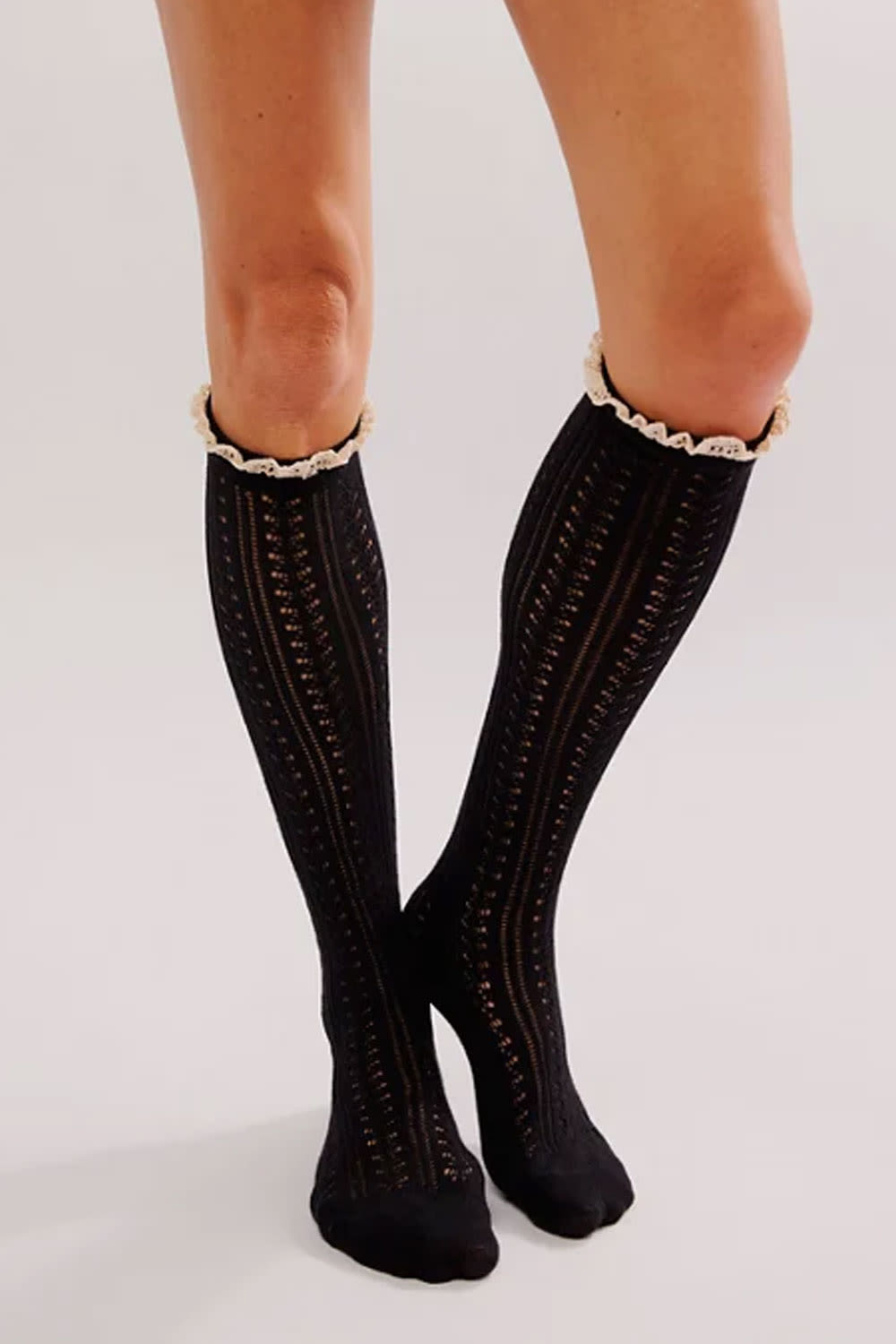 Black knee-high knit socks with a textured cable pattern and a cream ruffled trim at the top, shown on a model's legs against a light gray background.