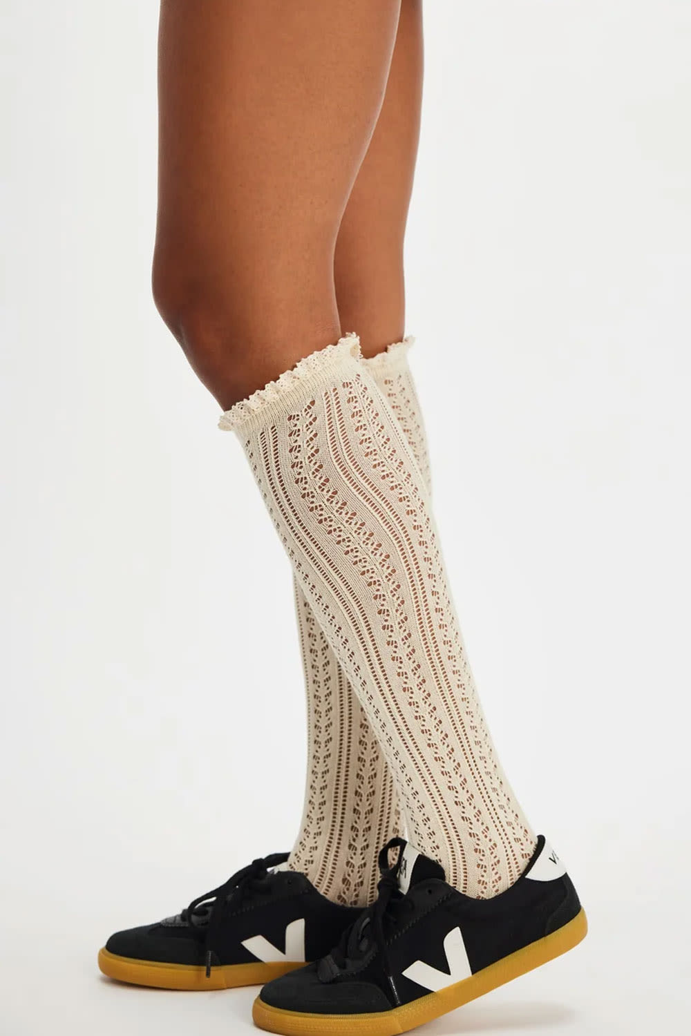 Cream cable-knit knee-high socks with a ruffled top edge, worn with black sneakers featuring white side stripes and yellow soles, shown on a model's legs against a white studio background.