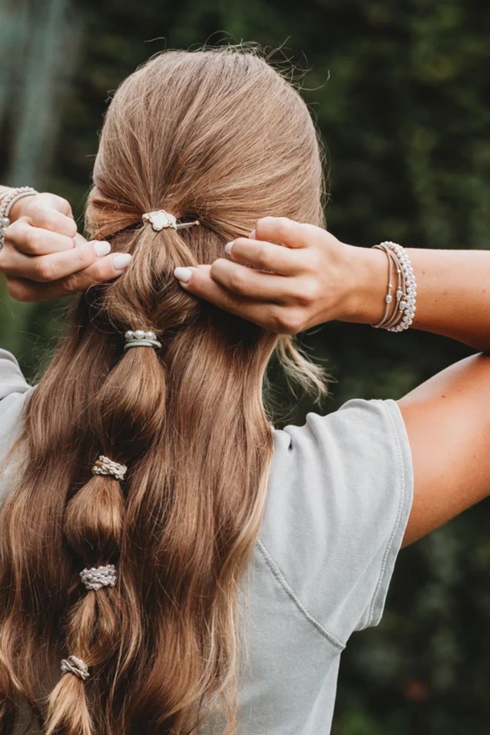 A woman viewed from behind with long light brown hair styled in a low braid, adjusting a small hair accessory with both hands. She is wearing a light gray T-shirt and multiple beaded bracelets, with a blurred green outdoor background.