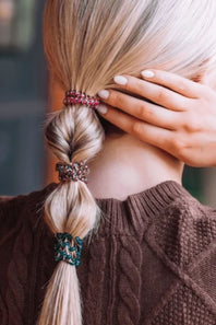 A close-up lifestyle shot of a blonde woman from behind, showcasing three decorative hair scrunchies in pink, brown, and teal holding her twisted ponytail. She is wearing a brown cable-knit sweater and touching her hair with one hand.