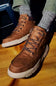 A pair of brown leather lace-up boots with chunky white soles, shown being worn with gray patterned socks on a wooden floor.