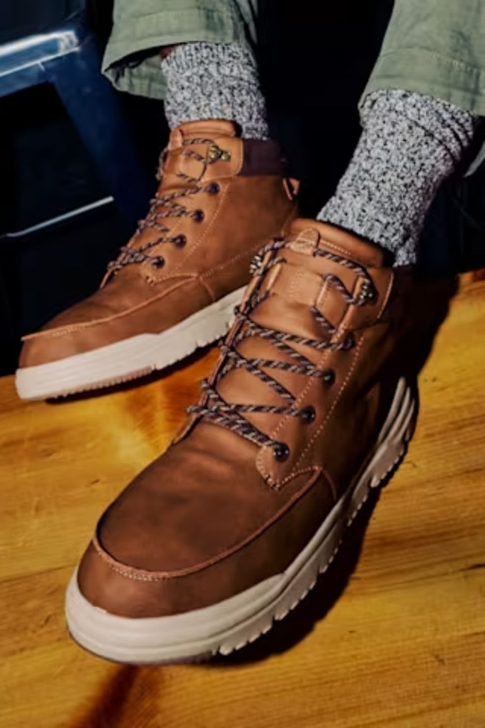 A pair of brown leather lace-up boots with chunky white soles, shown being worn with gray patterned socks on a wooden floor.