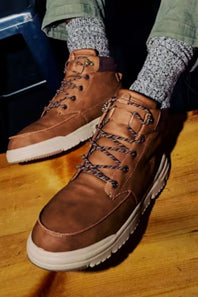 A pair of brown leather lace-up boots with chunky white soles, shown being worn with gray patterned socks on a wooden floor.