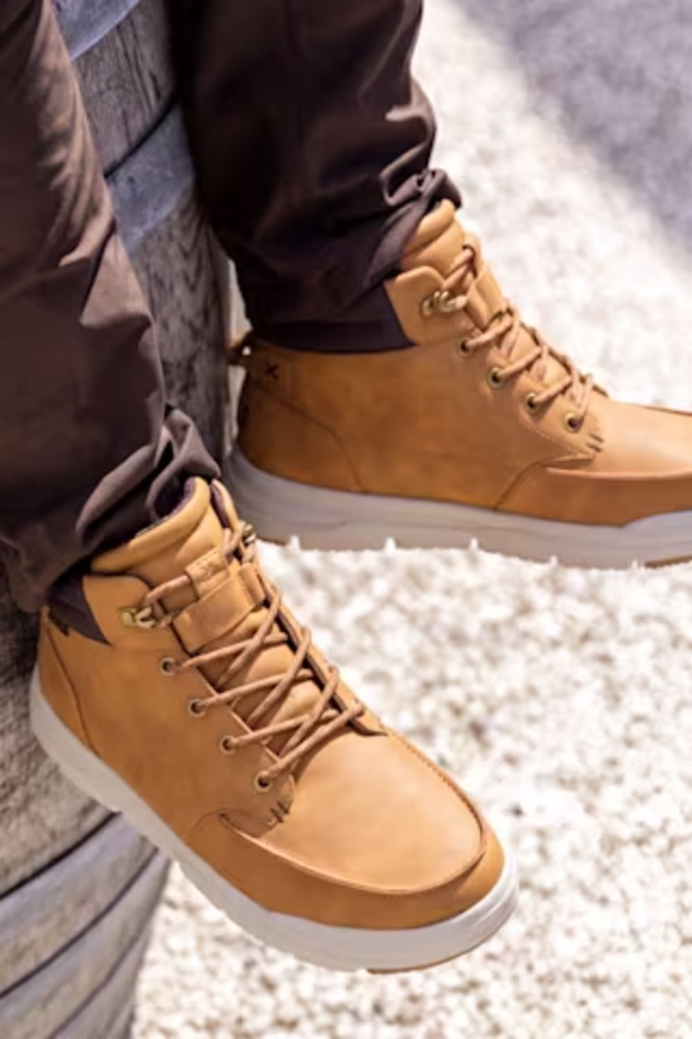 A close-up of a person wearing tan suede high-top lace-up boots with white soles, paired with dark brown pants, sitting on a textured surface.