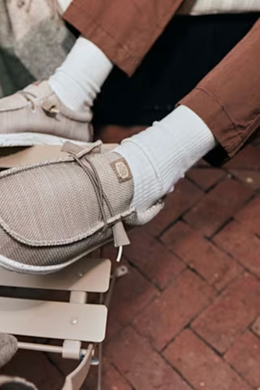 A person wearing beige slip-on lace-up shoes with white socks, sitting on a wooden bench on a brick-paved floor.