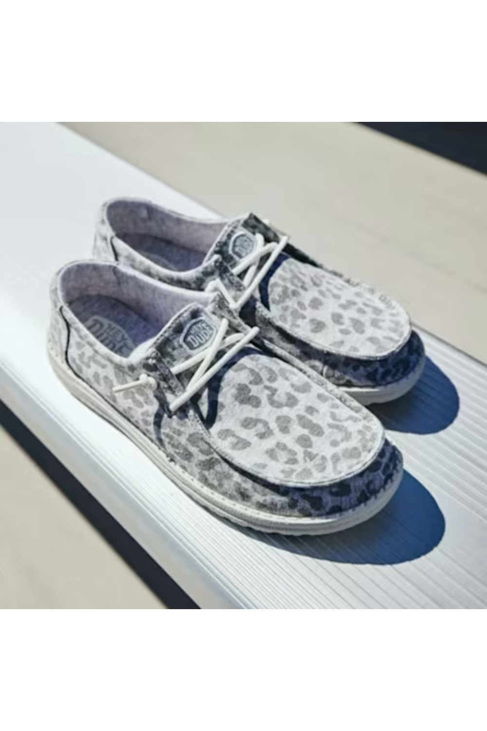 A pair of low-top boat-style shoes with a gray and white leopard print pattern, white laces, and dark blue side panels, displayed on a white textured surface.
