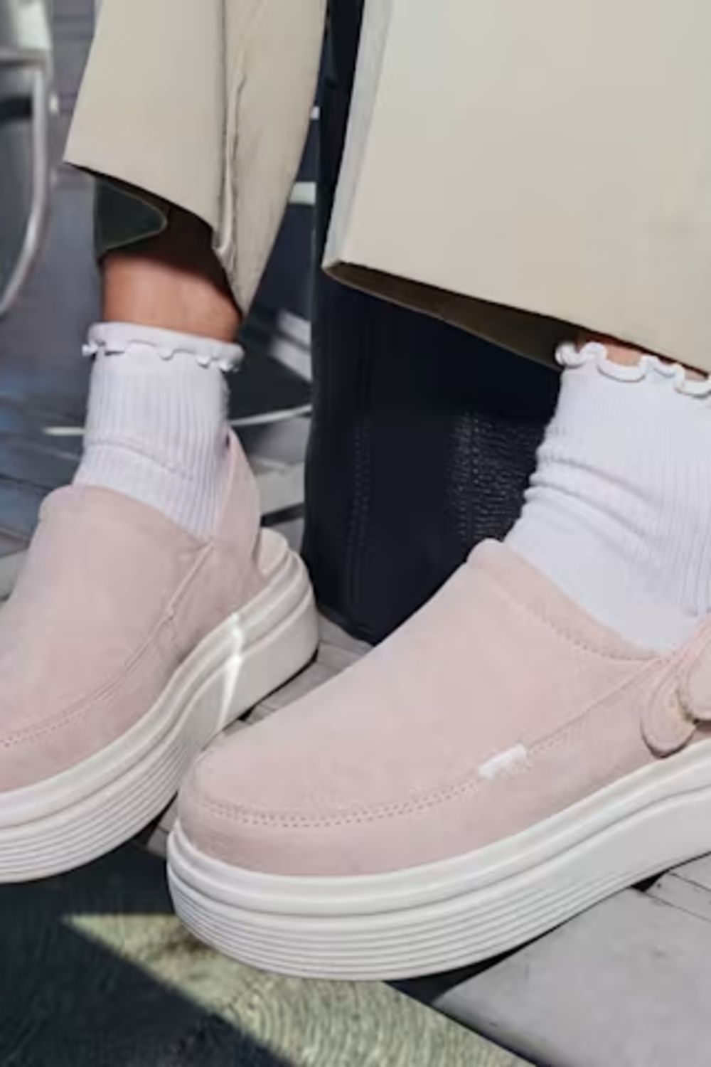 A person wearing a pair of light pink slip-on platform clogs with white ruffled socks and beige trousers, sitting on a bench.