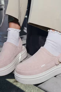 A person wearing a pair of light pink slip-on platform clogs with white ruffled socks and beige trousers, sitting on a bench.