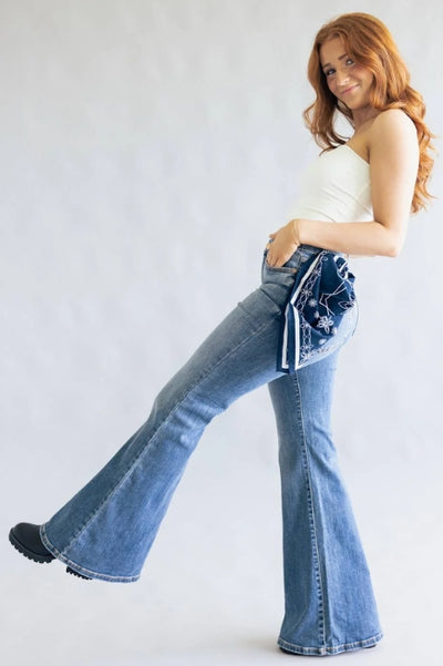 A model poses in profile wearing medium-wash high-rise flare jeans, a white strapless top, and black shoes, with a blue patterned bandana tucked into the back pocket against a neutral gray background.