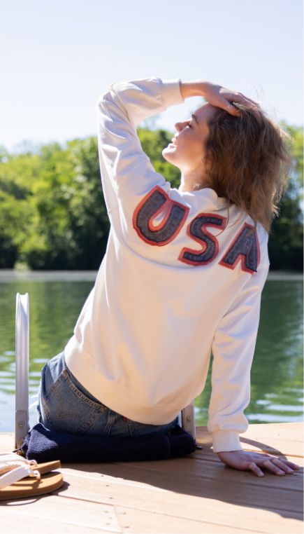 Girl siting on a lake deck facing the lake while leaning back on her right hand and left hand on her head wearing a white sweatshirt with "USA" across the top back