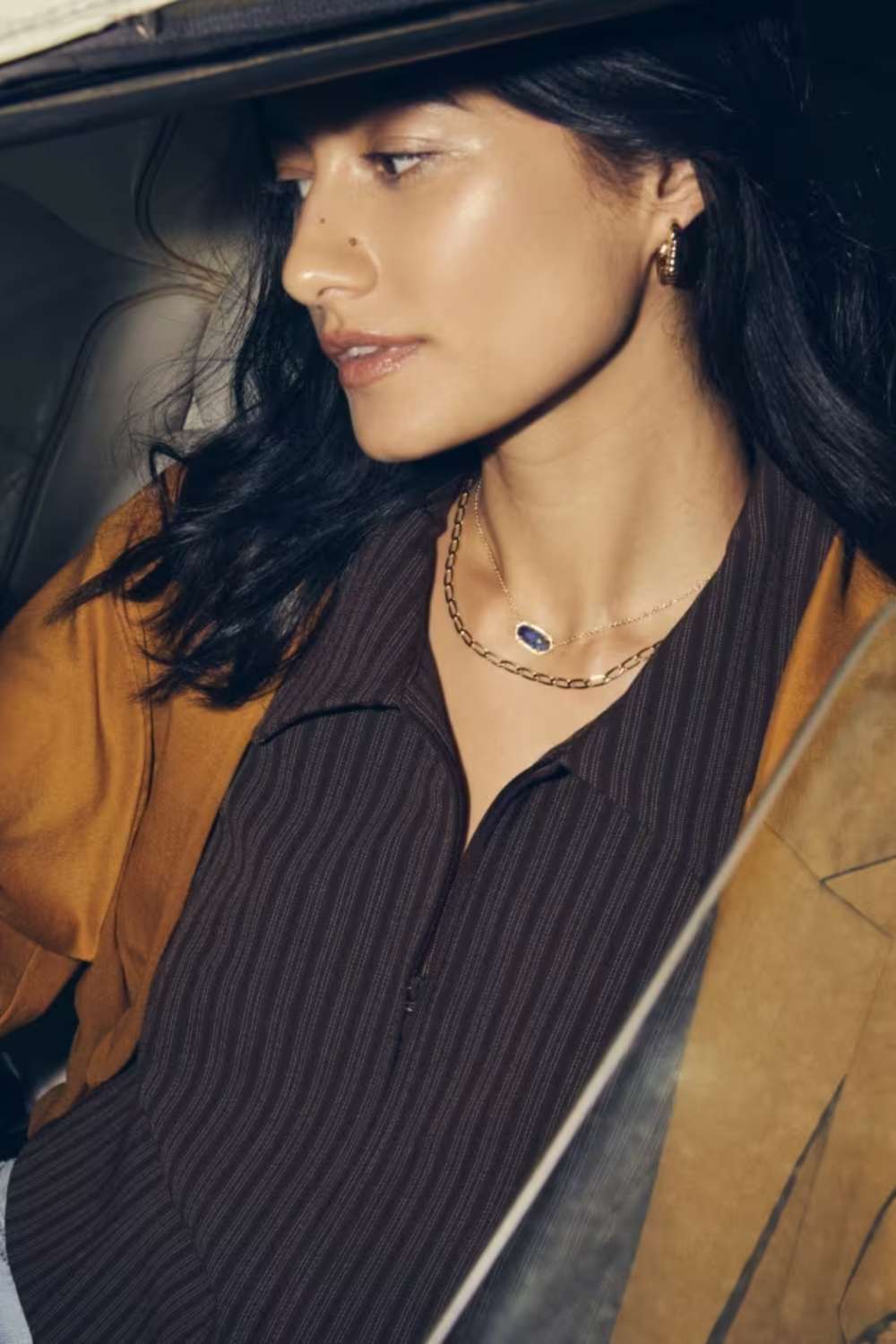 A close-up lifestyle shot of a woman wearing a gold chain necklace with a small oval blue stone pendant, styled with a dark brown pinstriped shirt and a mustard yellow jacket.
