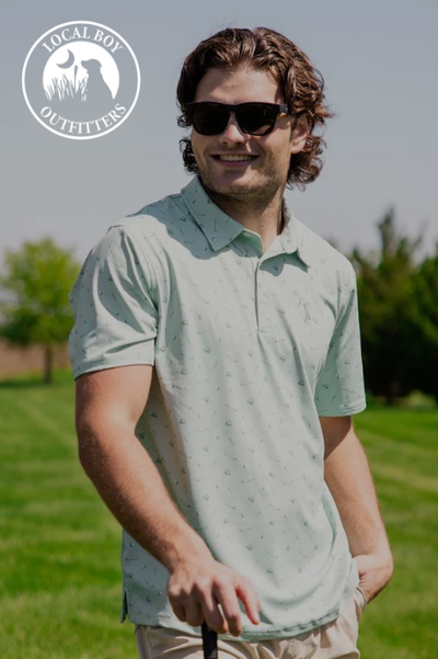 Smiling man wearing sunglasses and a light green polo stands outdoors on a sunny day, holding a golf club. Logo for 