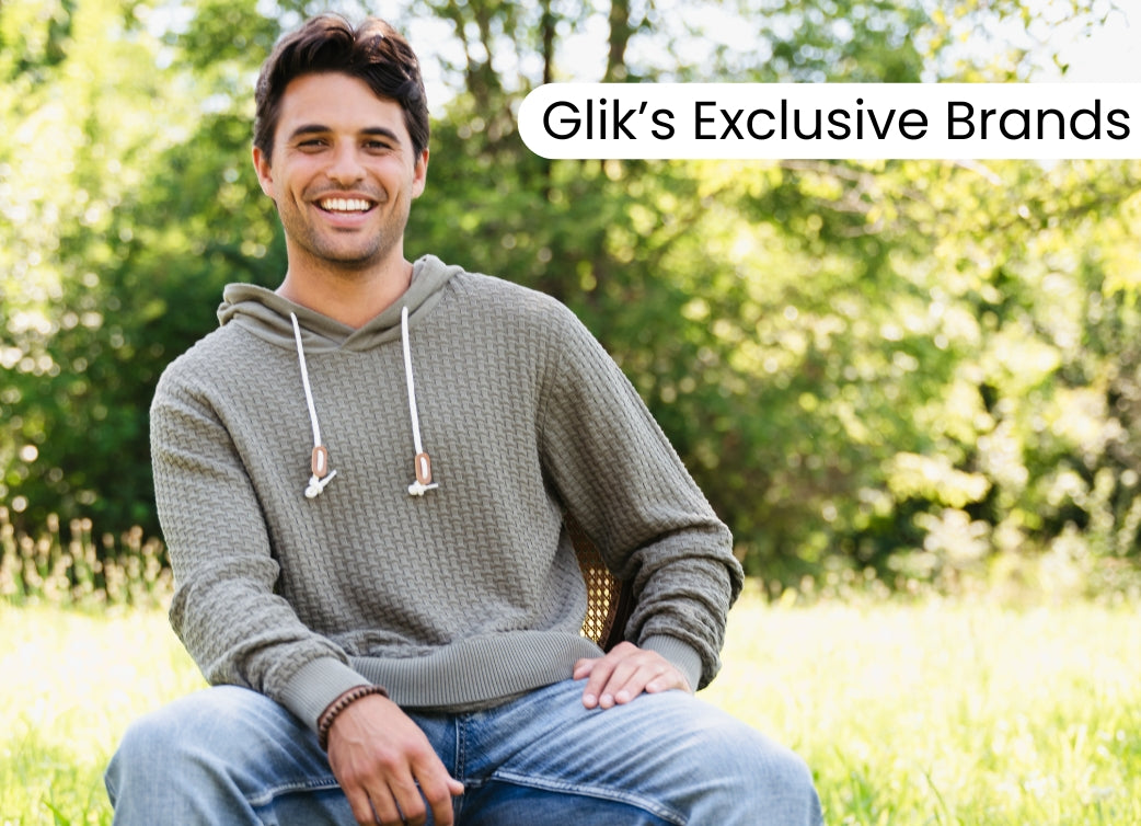 Promotional banner featuring a smiling man wearing a textured gray hoodie and blue jeans outdoors, with text reading “Glik’s Exclusive Brands” over a sunny wooded background.