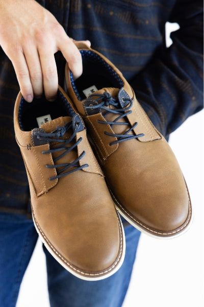 Person wearing a black long sleeve and blue jeans holding a pair of brown casual shoes with navy laces 