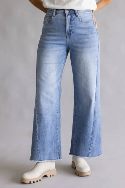 Front view of a model wearing light-wash high-rise cropped wide-leg jeans with raw hem edges, paired with white ankle boots against a neutral gray background.