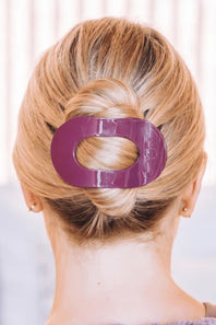 A close-up back view of a person with blonde hair styled in a low bun, secured with a glossy purple O-shaped hair claw clip.