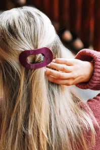 A close-up lifestyle shot of a person with long blonde hair wearing a large, dark purple plastic hair clip to secure a low ponytail. 