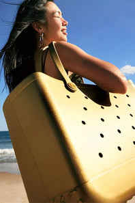 A woman on a beach carrying a large, structured tan tote bag with circular cutout details. The photo is taken from a low angle, showing the woman's profile and the bag against a bright blue sky and ocean background.