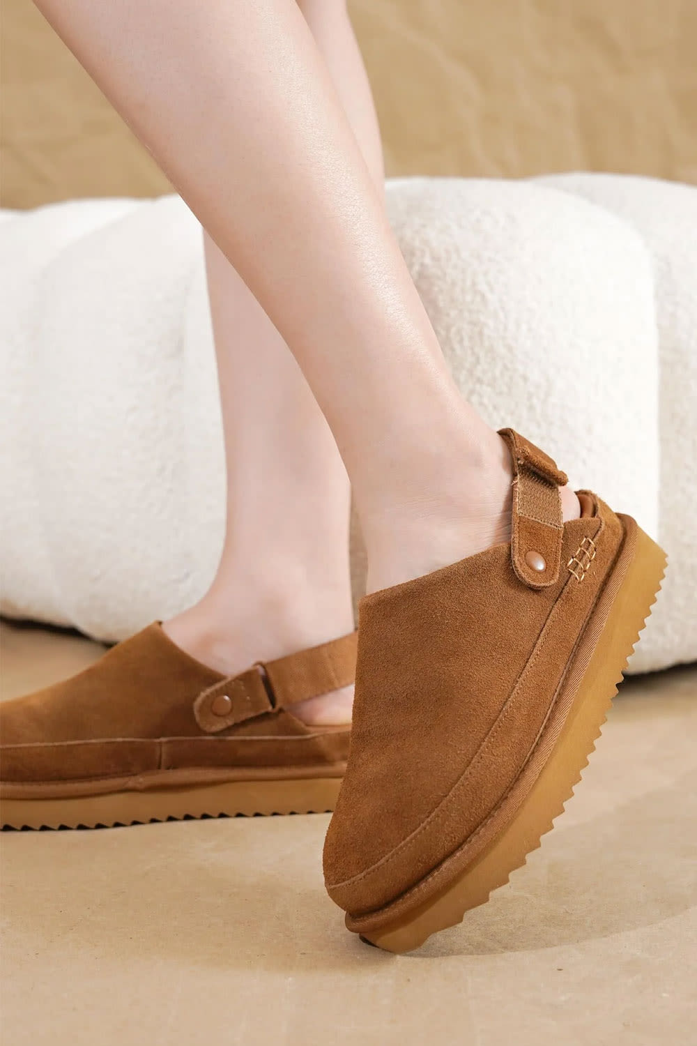 A pair of women's brown suede slip-on platform clog shoes with ankle straps, shown on a model's feet against a neutral beige and white background.