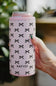 A hand holding a pink can cooler sleeve with a black bow pattern and the brand name 'KATYDID' printed on it, set against a blurred green and neutral background.