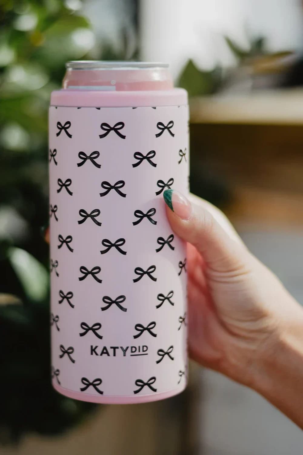 A hand holding a pink can cooler sleeve with a black bow pattern and the brand name 'KATYDID' printed on it, set against a blurred green and neutral background.