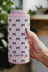 A hand holding a pink can cooler sleeve with a black bow pattern and the brand name 'KATYDID' printed on it, set against a blurred green and neutral background.