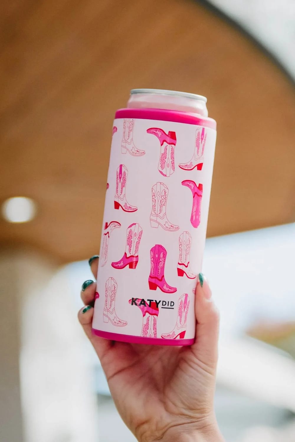 A hand holding a pink can cooler sleeve with a cowboy boot pattern, designed by Katydid.