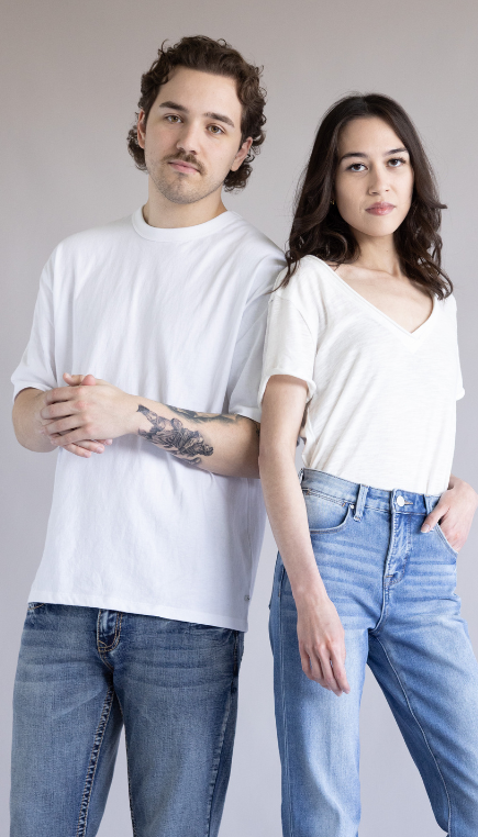 Two models stand side by side against a light gray background wearing white T-shirts and blue jeans. Male model wears a crewneck tee with medium-wash jeans, while the female model wears a V-neck tee with light-wash high-rise jeans.