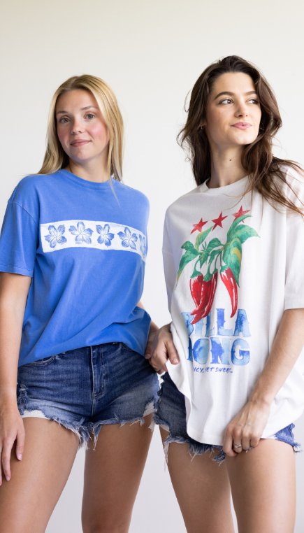 Two models stand side by side wearing graphic T-shirts and denim shorts against a light neutral background. One wears a blue T-shirt with a white floral stripe, while the other wears a white oversized T-shirt featuring a colorful chili pepper graphic and "BILLABONG" text.