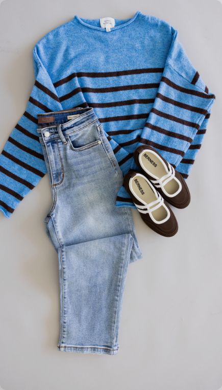 Flat lay of a casual outfit featuring a blue sweater with brown horizontal stripes, light wash jeans, and brown-and-white Mary Jane flats arranged on a light gray background.