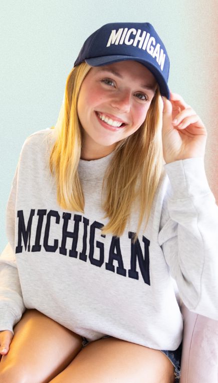Woman smiling while wearing a gray sweatshirt with "Michigan" across the front and a matching blue baseball cap embroidered with "Michigan."