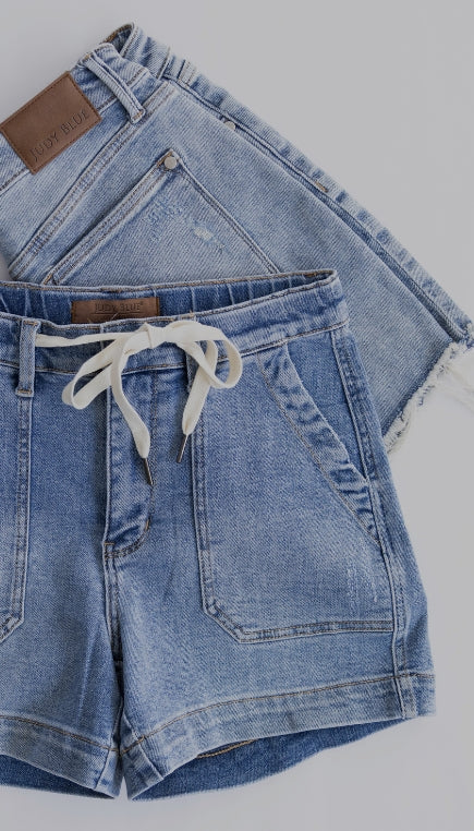 Flat lay of two light-wash denim bottoms arranged on a light gray background, featuring frayed-hem denim shorts and drawstring denim shorts with front patch pockets.