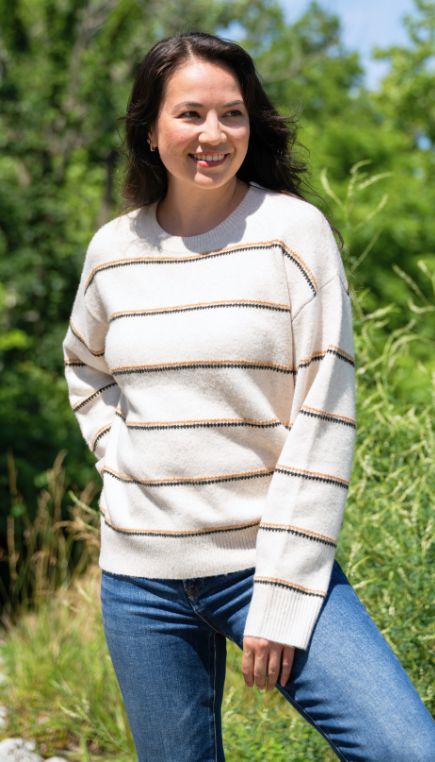 Woman standing outdoors, smiling, wearing a cream-colored sweater with thin brown stripes and blue jeans. Green foliage and trees are visible in the background under a clear sky.