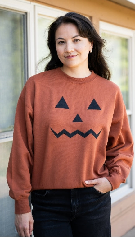 Woman wearing a jack-o-lantern face rust colored crewneck sweatshirt and black jeans standing in front of some building windows