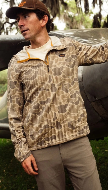 Man standing underneath some trees wearing a brown colored camo quarter zip, dark colored pants and a brown hat