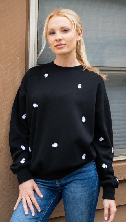 Women standing in front of brown wall and window wearing a black crewneck sweatshirt with white embroidered ghosts and dark wash denim jeans