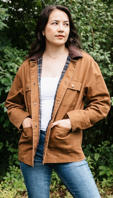 Woman standing outdoors in front of greenery wearing a brown barn jacket with a white t shirt layered underneath and blue denim jeans