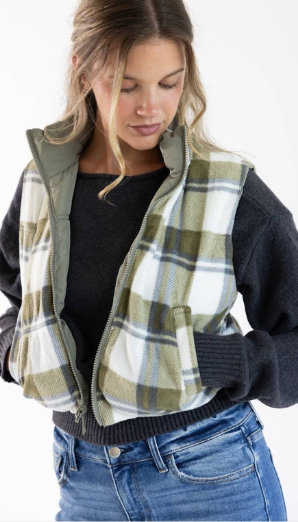 A woman wearing a olive green and white plaid puffer vest with a charcoal sweater layered underneath with denim jeans on a white background