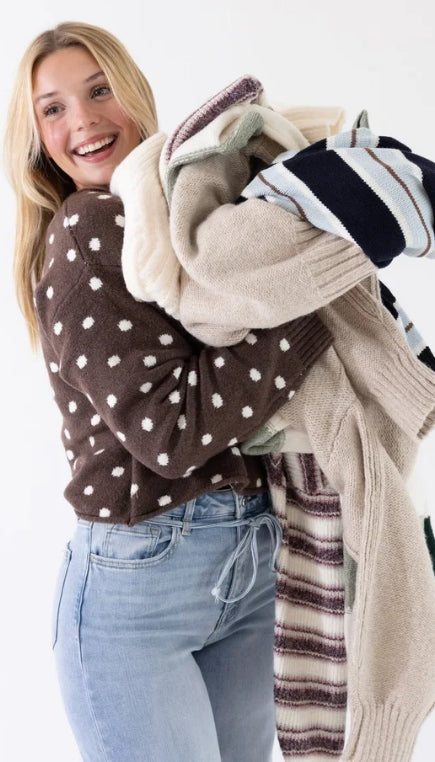 Woman wearing a brown sweater with white polka dot and denim jeans holding multiple different sweaters in front of a white background