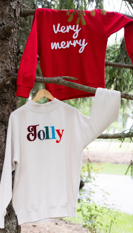 Two sweatshirt hanging on branches of a tree. One sweatshirt is red with text Very Merry, the other sweatshirt is beige with multi colored text Jolly