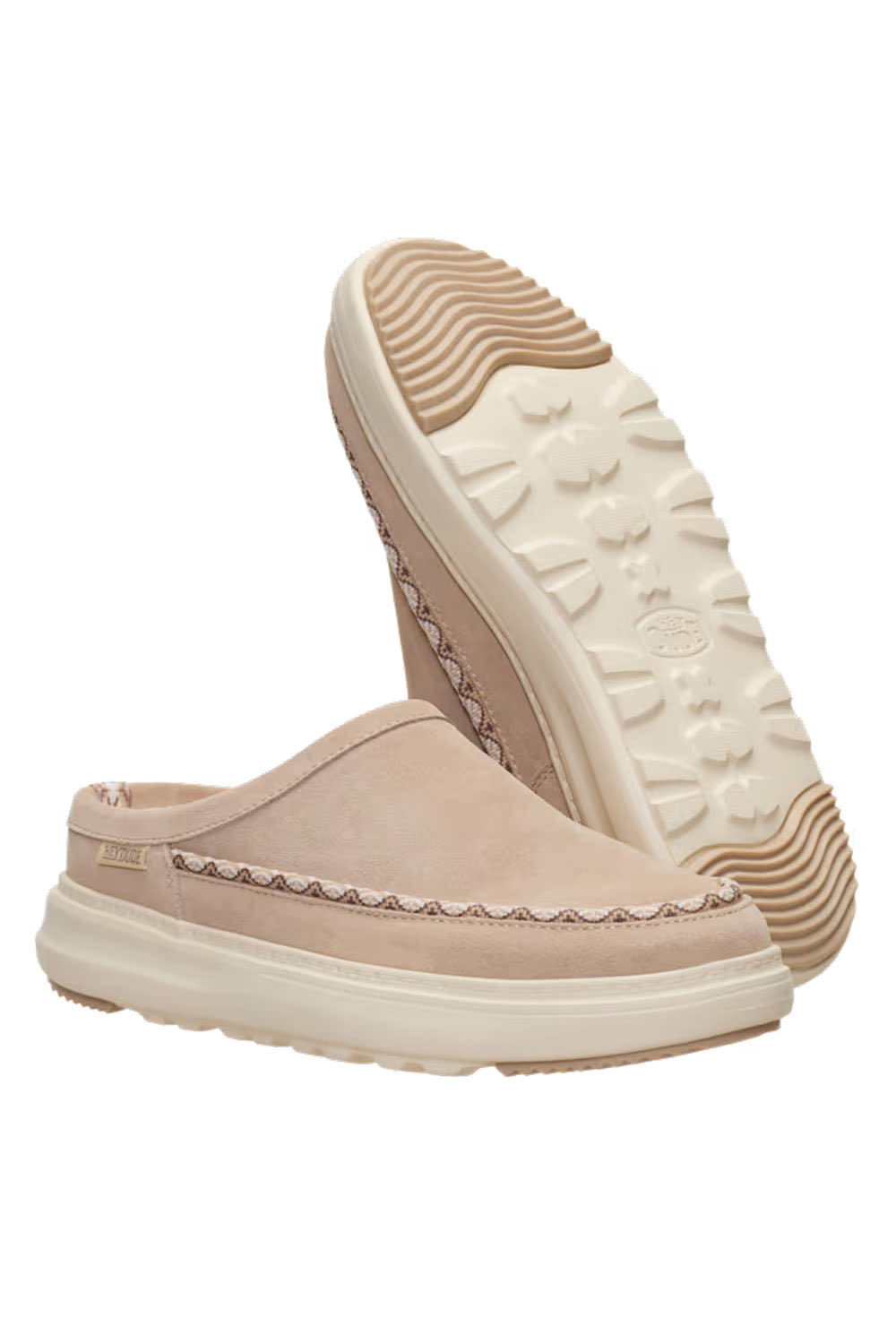 A pair of slip-on clog-style shoes in a light beige suede-like material, with one shoe standing upright to show the textured off-white sole and the other lying flat to show the side profile and decorative stitching.