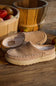 A pair of tan slip-on slippers with cream lining and decorative stitching, displayed on a rustic wooden surface with baskets of apples in the background. The brand name HEY DUDE