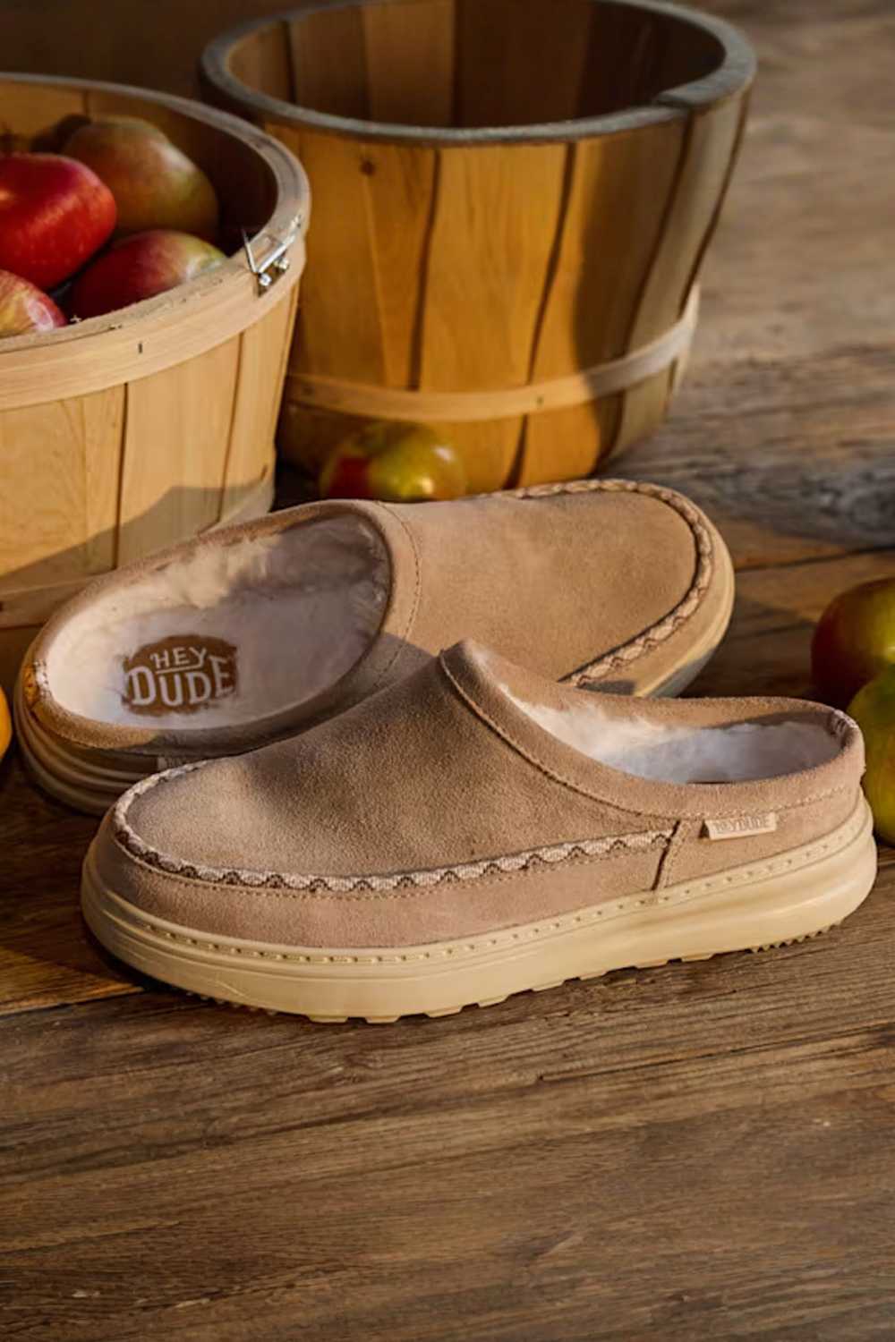 A pair of tan slip-on slippers with cream lining and decorative stitching, displayed on a rustic wooden surface with baskets of apples in the background. The brand name HEY DUDE
