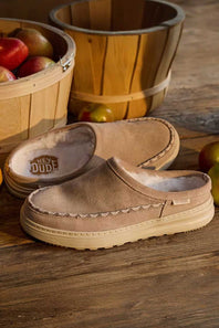A pair of tan slip-on slippers with cream lining and decorative stitching, displayed on a rustic wooden surface with baskets of apples in the background. The brand name HEY DUDE