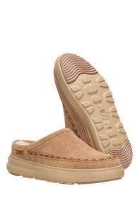 A pair of tan suede clogs with thick beige soles, shown against a plain white background. One clog is positioned upright to show the side profile, while the other is turned upside down to display the treaded outsole.