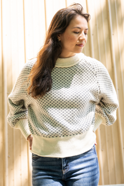 Women standing in front of tan wall with sun reflections wearing a white and grey diamond pattern sweater and blue jeans