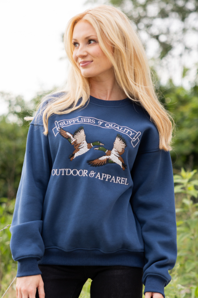 Women standing outdoors wearing a navy blue sweatshirt with mallard ducks and reads SUPPLIERS & QUALITY OUTDOR & APPAREL and black pants