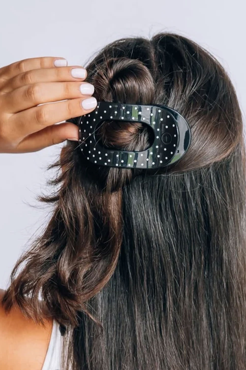 A close-up of a woman's hair styled with a half-up bun, wearing a black hair claw clip with white polka dots. A hand with white manicured nails is adjusting the clip.
