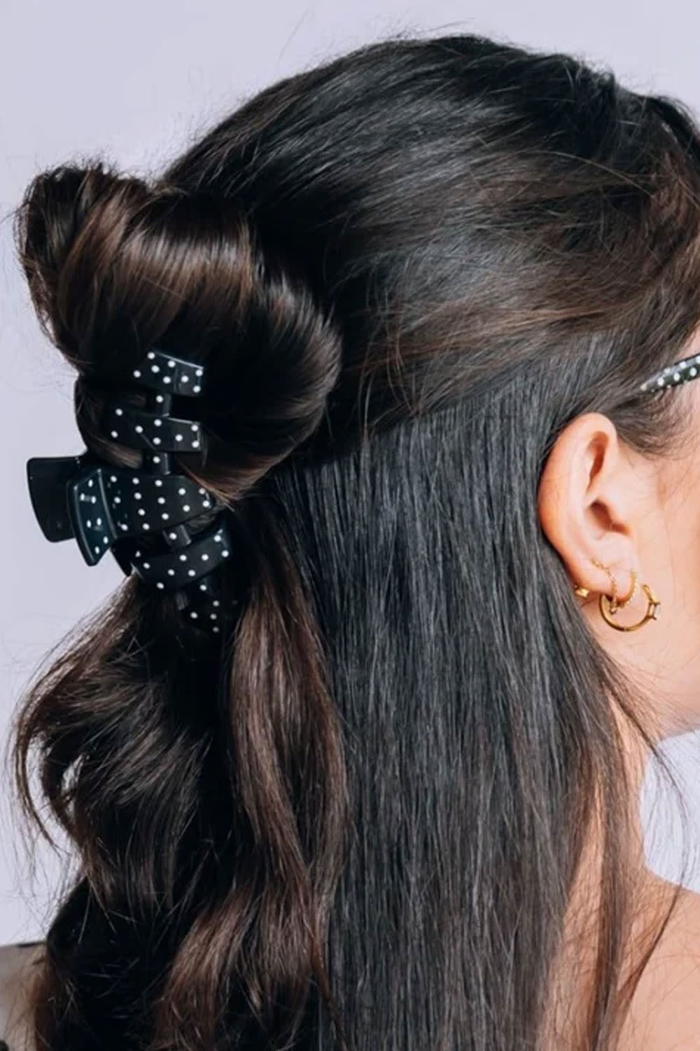 A close-up side view of a person with long dark brown hair, featuring a black hair claw clip with white polka dots holding a half-up bun.